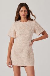 A woman is wearing a short, beige textured dress with short sleeves. The dress has a round neckline and a slightly loose fit, ending above the knees. The fabric appears to be a woven or boucle material with a multicolored pattern in light shades.
